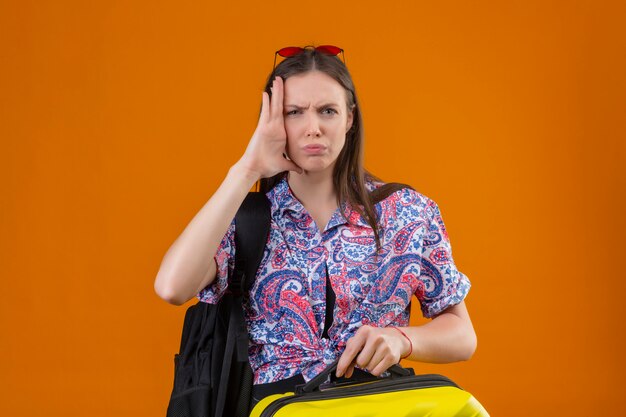 Femme jeune voyageur dérangé portant des lunettes de soleil rouges sur la tête debout avec sac à dos tenant la valise à la tête de contact stressée et ennuyée avec la main sur l'orange
