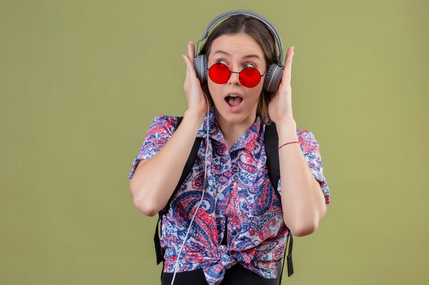 Femme jeune voyageur portant des lunettes de soleil rouges et avec sac à dos, écouter de la musique à l'aide d'un casque à la surprise sur mur vert