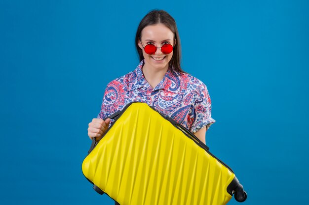 Femme jeune voyageur portant des lunettes de soleil rouges tenant une valise jaune à la recherche de sourire heureux et positif joyeusement sur mur bleu