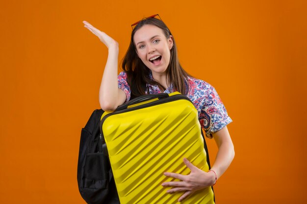 Femme jeune voyageur portant des lunettes de soleil rouges sur la tête debout avec sac à dos tenant valise regardant la caméra et présentant avec le bras de la main quelque chose de souriant avec un visage heureux sur backg orange