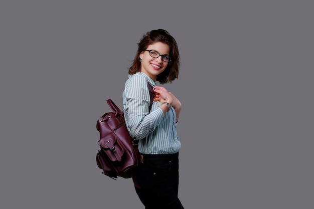 Femme jeune voyageur positif avec sac à dos en cuir isolé sur fond gris.