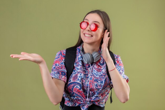 Femme jeune voyageur avec sac à dos et casque portant des lunettes de soleil rouges souriant joyeusement présentant avec bras de main debout sur mur vert