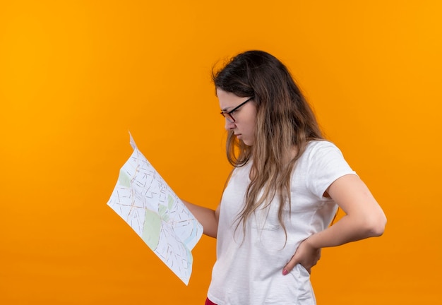 Femme jeune voyageur en t-shirt blanc tenant la carte en la regardant avec une expression pensive sur le visage pensant debout sur un mur orange