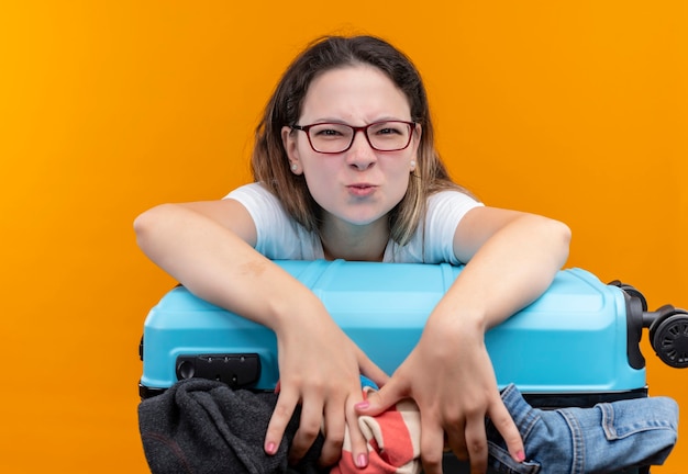 Femme jeune voyageur en t-shirt blanc tenant une valise pleine de vêtements en fronçant les sourcils à la recherche debout