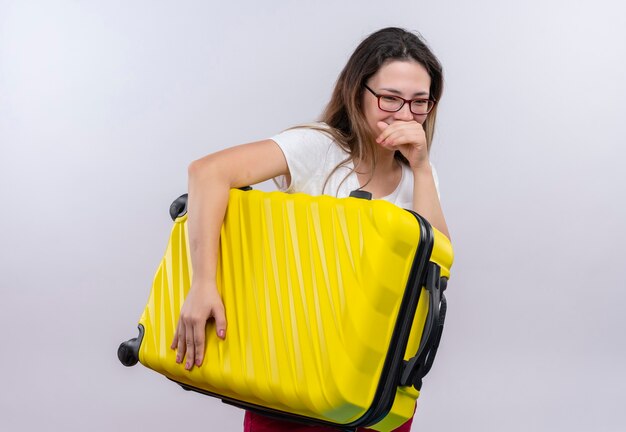 Femme jeune voyageur en t-shirt blanc tenant valise en riant heureux et positif debout sur un mur blanc