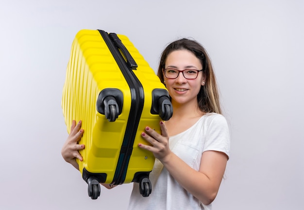 Femme jeune voyageur en t-shirt blanc tenant valise souriant heureux et positif debout sur un mur blanc