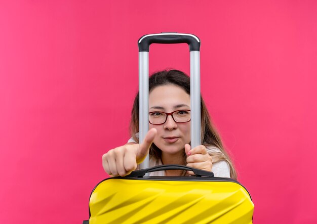 Femme jeune voyageur en t-shirt blanc tenant une valise de voyage montrant les pouces vers le haut à la confiance debout sur le mur rose