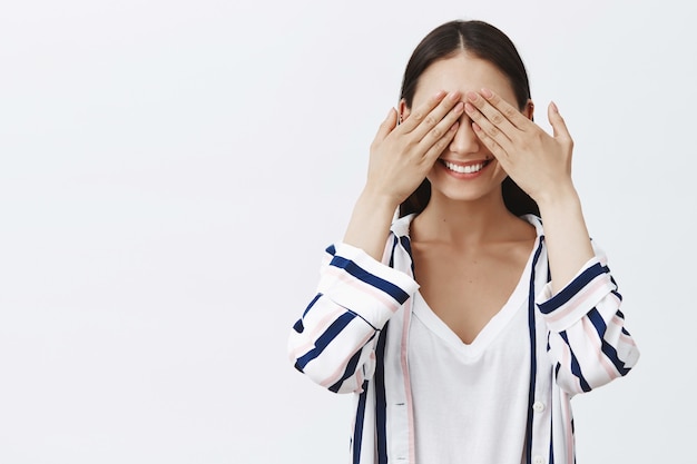 femme jouant à cache-cache. Ravie de belle femme élégante en chemisier rayé, couvrant les yeux avec des paumes et souriant avec une expression intriguée, en attente de surprise