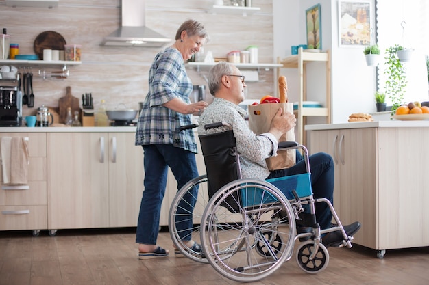 Femme joyeuse aidant son mari handicapé dans la cuisine. Femme âgée prenant un sac en papier d'épicerie d'un mari handicapé en fauteuil roulant. Personnes mûres avec des légumes frais du marché. Vivre avec un handicap