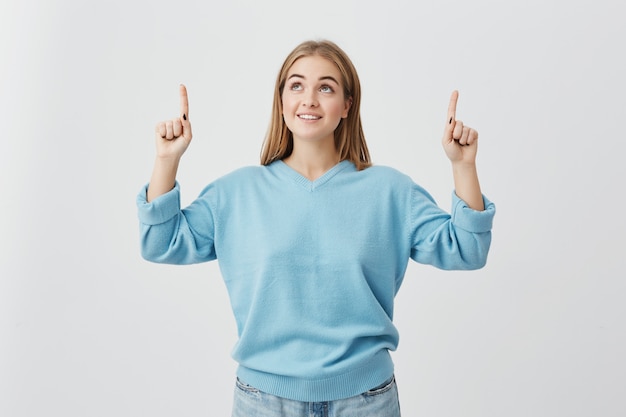 Femme joyeuse aux cheveux blonds, portant un pull bleu et un jean, pointant vers le haut avec ses doigts étant heureux de voir quelque chose. Fille aux cheveux blonds souriant joyeusement avec des dents