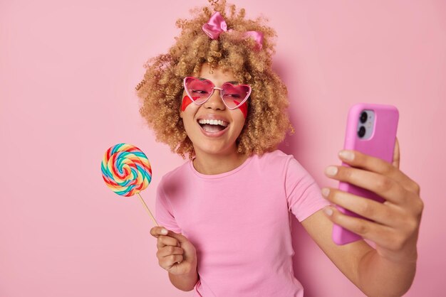 Une femme joyeuse aux cheveux bouclés tient de gros bonbons au caramel arc-en-ciel sur un bâton rit avec joie prend un selfie via un smartphone insensé autour de porte des lunettes de soleil et un t-shirt décontracté isolé sur fond rose