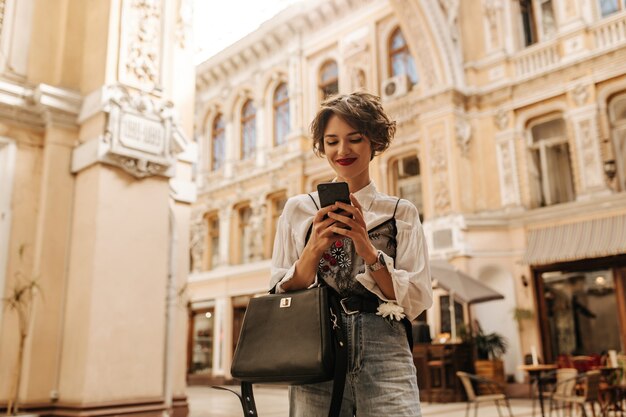 Femme joyeuse aux cheveux courts tenant le téléphone dans la rue. Femme élégante en chemise et jeans avec sac à main noir souriant en ville.