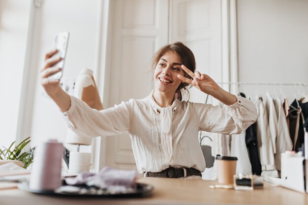 Femme joyeuse en chemisier élégant prenant selfie et montrant un signe de paix