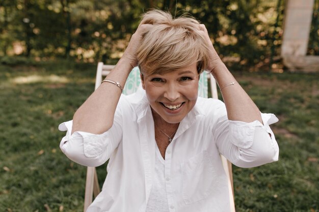 Femme joyeuse avec une coiffure à la mode blonde et des accessoires en chemise blanche élégante riant et regardant dans la caméra dans le parc