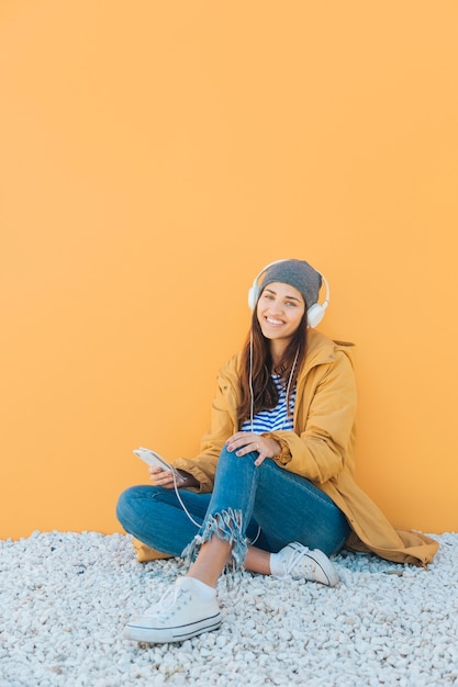 femme joyeuse écoute la musique sur un téléphone intelligent assis sur un tapis contre une surface jaune