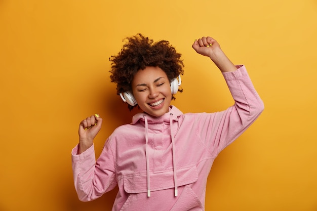 Une femme joyeuse et expressive écoute de la musique dans des écouteurs, aime des mélodies agréables, a de la bonne humeur, danse sans soucis, sourit largement, porte un sweat-shirt rose, pose contre un mur jaune. Gens, loisirs