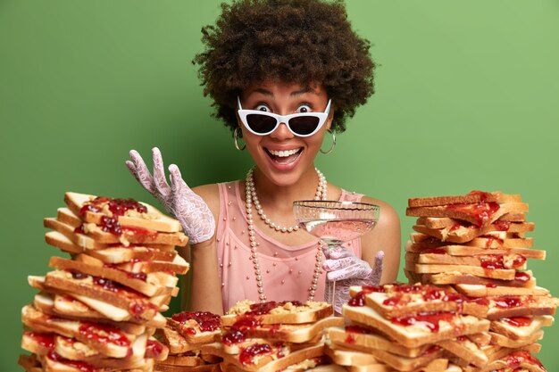 Une femme joyeuse à la peau sombre et aux cheveux bouclés, vêtue de vêtements élégants, porte des lunettes de soleil, boit un cocktail alcoolisé, entend d'excellentes nouvelles de l'interlocuteur, se tient près de la pile de sandwichs.