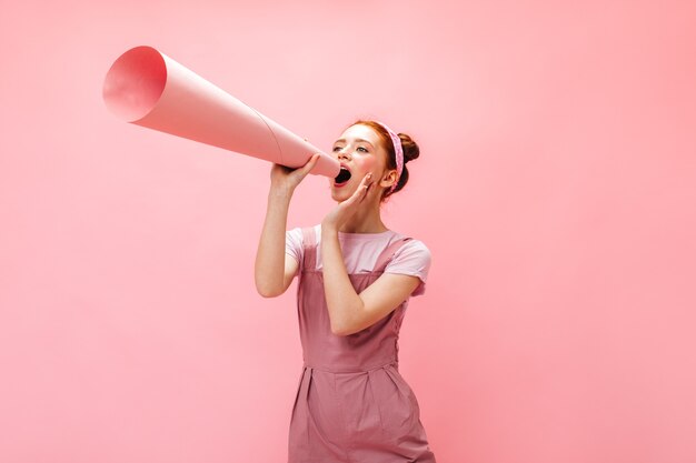 Femme joyeuse avec des petits pains hurle dans l'embout buccal. femme en salopette posant sur fond rose.