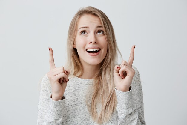 Femme joyeuse portant des cheveux blonds lâches en regardant les doigts pointés vers le haut à l'espace de copie au-dessus de la tête.