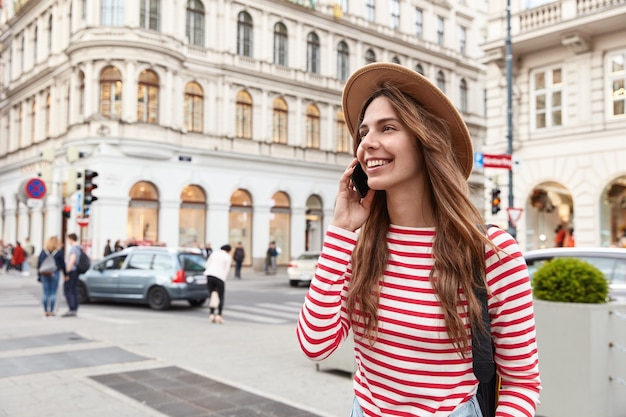 Photo gratuite une femme joyeuse se promène dans la ville, aime la communication, garde un smartphone moderne près de l'oreille, concentré de côté