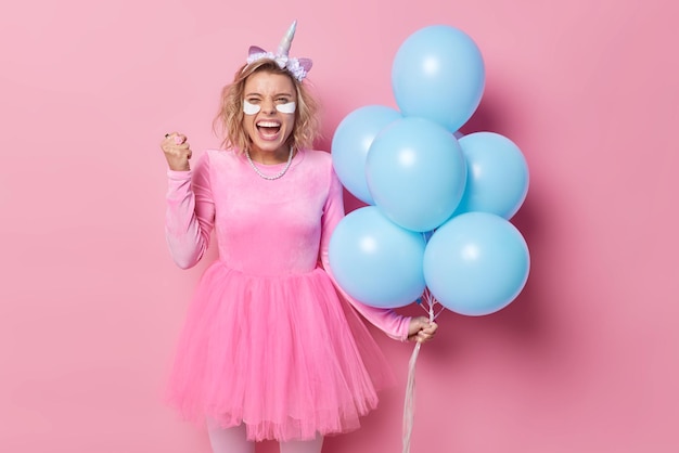 Photo gratuite une femme joyeuse serre le poing célèbre une occasion spéciale sent que le succès porte une robe de fête tient un tas de ballons à l'hélium bleu applique des patchs de beauté sous les yeux pour éliminer les rides pose à l'intérieur