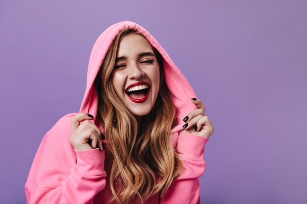 Femme joyeuse en sweat à capuche rose riant sur un mur isolé