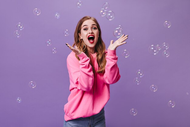 Femme joyeuse en sweat à capuche rose surprise en regardant dans la caméra et en jouant avec des bulles