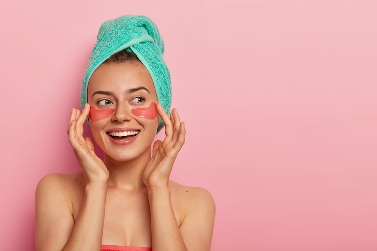 Photo gratuit une femme joyeuse touche doucement le visage, sourit agréablement, applique des patchs d'hydrogel sous les yeux, se tient nue, a une peau saine
