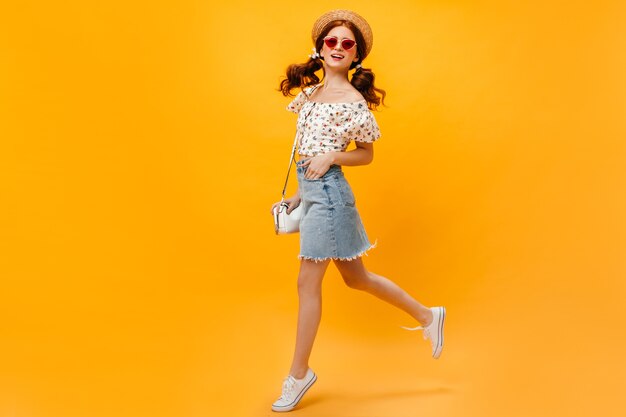 femme en jupe en jean, T-shirt blanc et canotier sautant sur fond orange. Femme à lunettes de soleil souriant.
