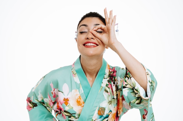 Femme en kimono japonais traditionnel faisant signe ok regardant à travers les doigts heureux et joyeux souriant sur blanc
