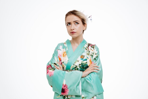 Femme en kimono japonais traditionnel levant avec une expression pensive avec les bras croisés pensant sur le blanc