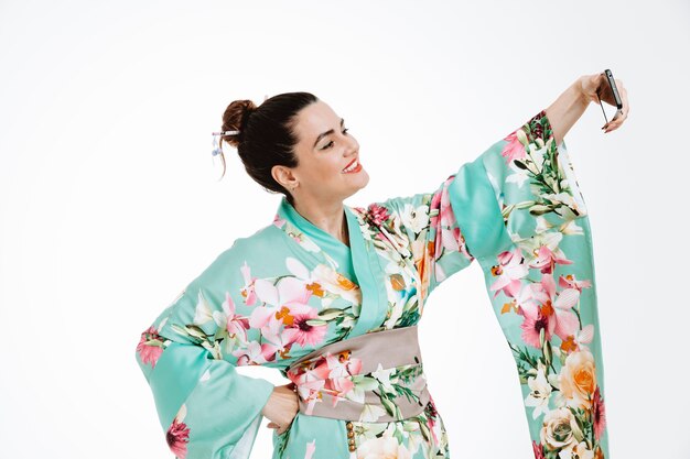 Femme en kimono japonais traditionnel souriant heureux et positif faisant gaiement selfie à l'aide de smartphone sur blanc