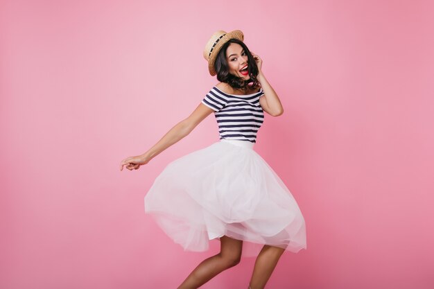 Femme latine drôle en chapeau de paille exprimant le bonheur et la danse. Modèle féminin émotionnel en jupe blanche posant