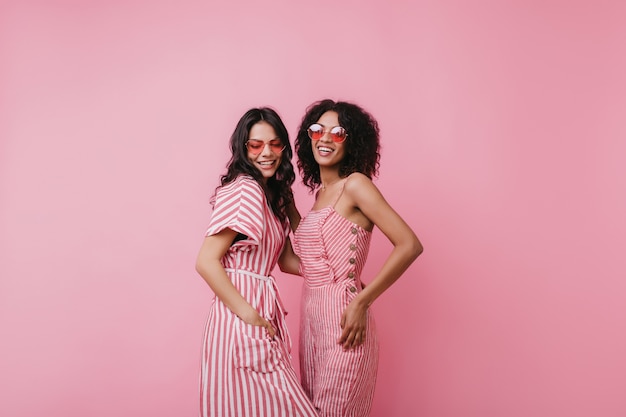 Femme latine galbée en tenue rayée posant avec une amie africaine. Photo intérieure de deux jolies femmes portant des lunettes de soleil roses à la mode.