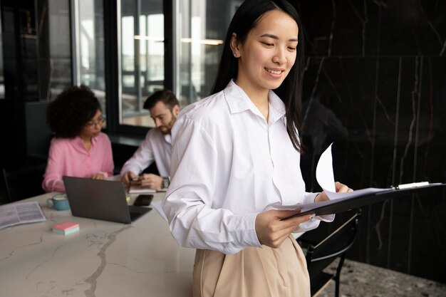 Femme lisant des documents pour le travail pendant que ses collègues utilisent un ordinateur portable