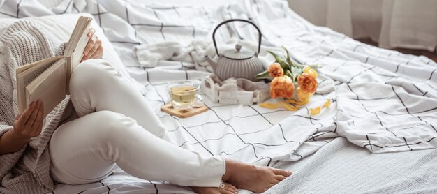 Femme lit un livre au lit avec du thé et un bouquet de tulipes