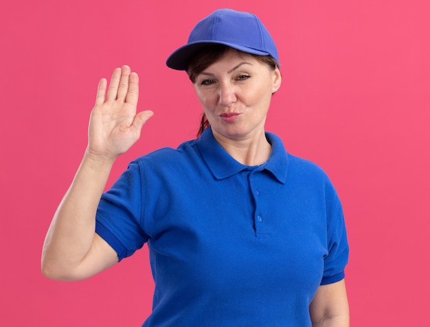 Photo gratuite femme de livraison d'âge moyen en uniforme bleu et cap à l'avant avec une expression confiante montrant la main ouverte debout sur le mur rose