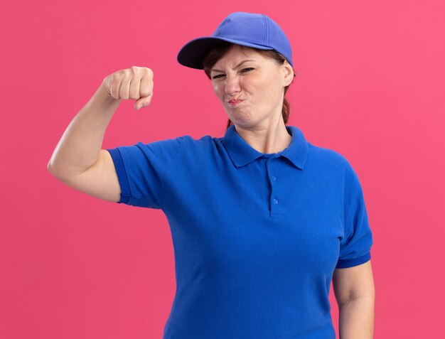 Femme de livraison d'âge moyen en uniforme bleu et cap à l'avant montrant le poing fermé d'être mécontent debout sur le mur rose