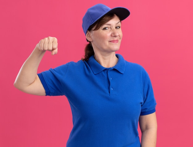 Femme de livraison d'âge moyen en uniforme bleu et cap à l'avant montrant le poing fermé souriant confiant debout sur le mur rose