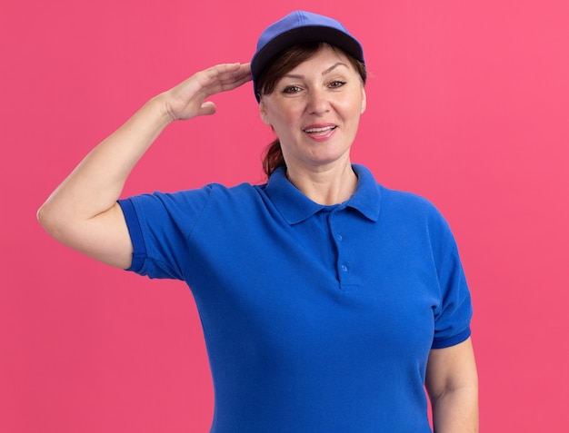 Femme de livraison d'âge moyen en uniforme bleu et cap à l'avant souriant confiant saluant debout sur le mur rose