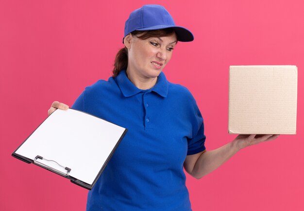 Femme de livraison d'âge moyen en uniforme bleu et cap holding boîte en carton et presse-papiers avec des pages blanches à la confusion et mécontent debout sur le mur rose