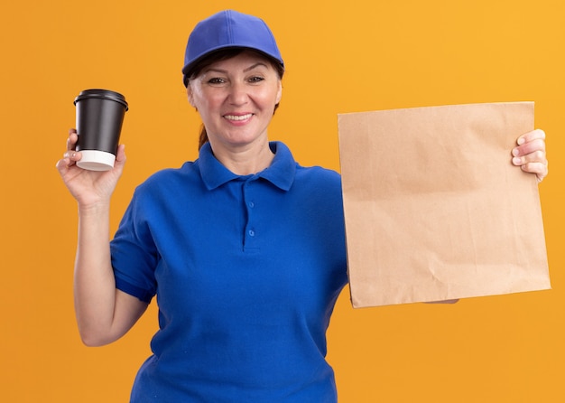 Femme de livraison d'âge moyen en uniforme bleu et cap holding paper package montrant tasse de café à l'avant avec sourire sur le visage debout sur le mur orange