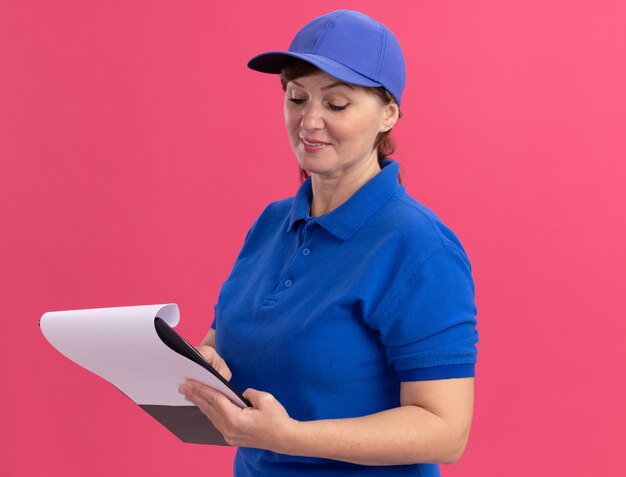 Femme de livraison d'âge moyen en uniforme bleu et cap holding presse-papiers avec des pages vierges à la regarder avec le sourire sur le visage debout sur le mur rose