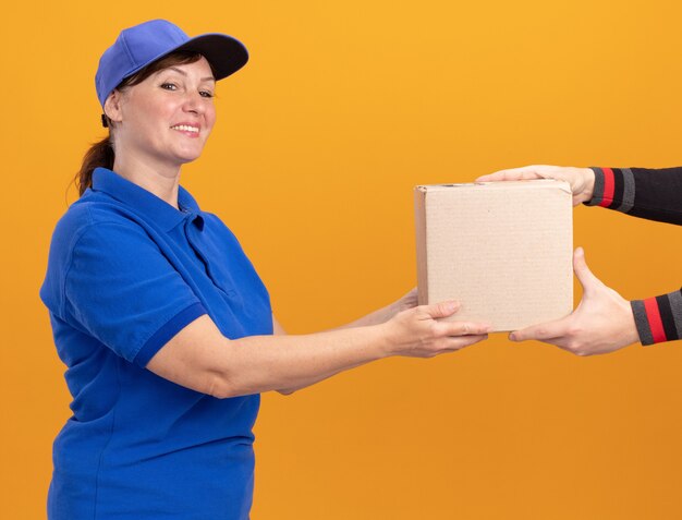 Femme de livraison d'âge moyen en uniforme bleu et capuchon donnant une boîte en carton à un client souriant debout sur un mur orange