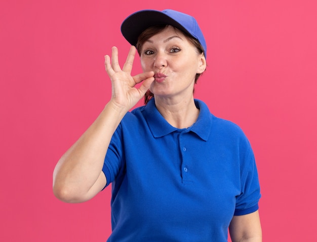 Femme de livraison d'âge moyen en uniforme bleu et casquette faisant un geste de silence comme la fermeture de la bouche avec une fermeture à glissière debout sur un mur rose