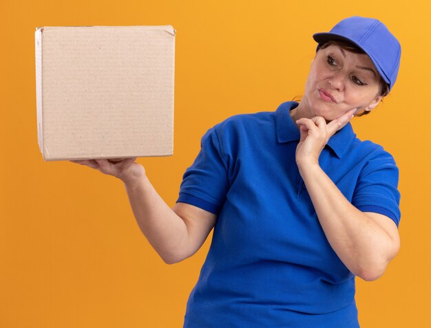 Femme de livraison d'âge moyen en uniforme bleu et casquette tenant une boîte en carton à la regarder perplexe debout sur un mur orange