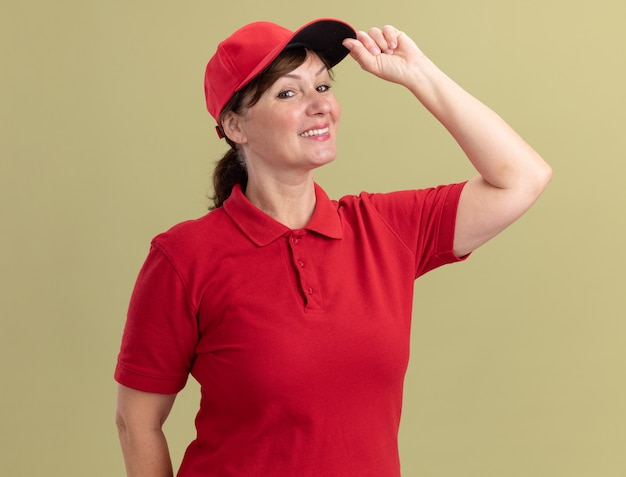 Photo gratuite femme de livraison d'âge moyen en uniforme rouge et cap à l'avant souriant confiant toucher sa casquette debout sur mur vert