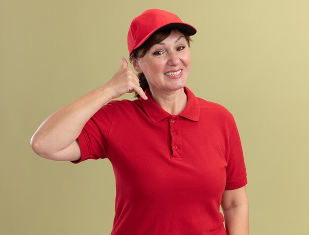 Femme de livraison d'âge moyen en uniforme rouge et cap à l'avant souriant faisant appelez-moi geste debout sur mur vert