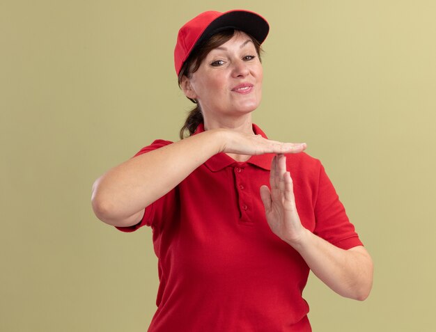 Femme de livraison d'âge moyen en uniforme rouge et cap à l'avant souriant faisant le geste du temps avec les mains debout sur le mur vert