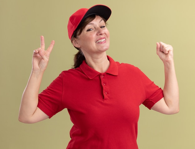Femme de livraison d'âge moyen en uniforme rouge et cap à l'avant souriant avec visage heureux montrant v-sign et levant le poing debout sur le mur vert